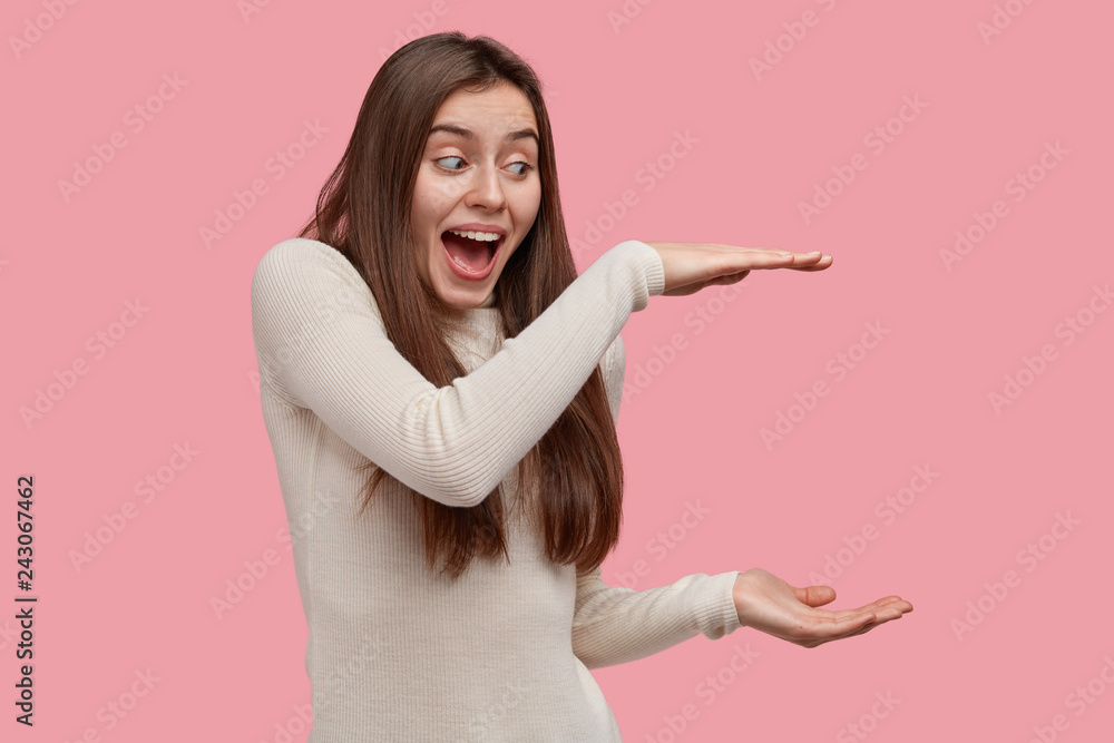 Happy overemotive young woman shows big size of recieved present, shares positive emotions with friend, shapes height of gift box with both hands, wears casual turtleneck sweater, models indoor Happy overemotive young woman shows big size of recieved present, shares positive emotions with friend, shapes height of gift box with both hands, wears casual turtleneck sweater, models indoor
