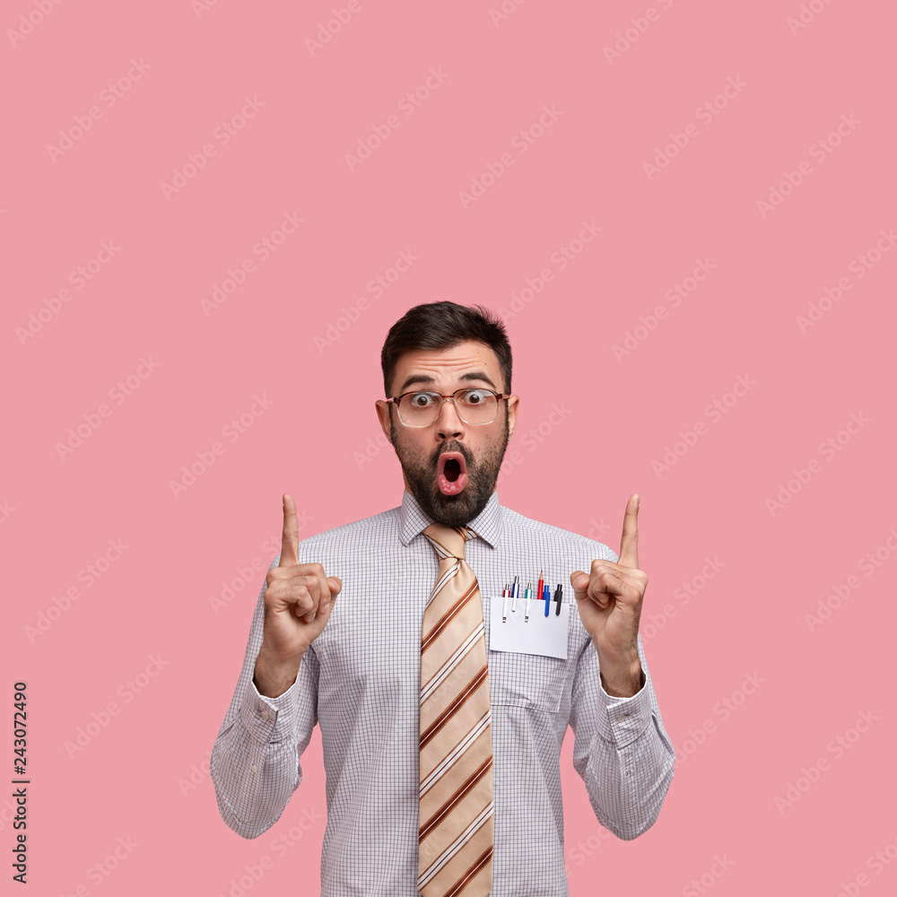 Indoor shot of surprised young entrepreneur points with both index fingers upwards, shows blank space, wears formal shirt and tie, isolated agaist pink background, looks concerned with disbelief.
