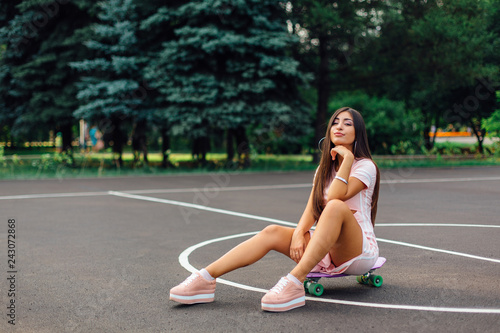 Portrait of a smiling charming brunette female sitting on her skateboard on a basketball court.
