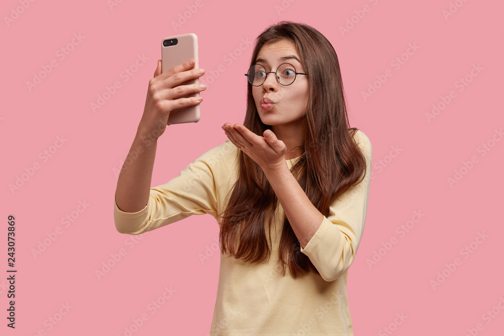 Pretty Caucasian woman makes air kiss at camera of cell phone, has video call with boyfriend on distance, flirts and says goodbye, wears round spectacles and yellow sweater, isolated on pink wall Pretty Caucasian woman makes air kiss at camera of cell phone, has video call with boyfriend on distance, flirts and says goodbye, wears round spectacles and yellow sweater, isolated on pink wall