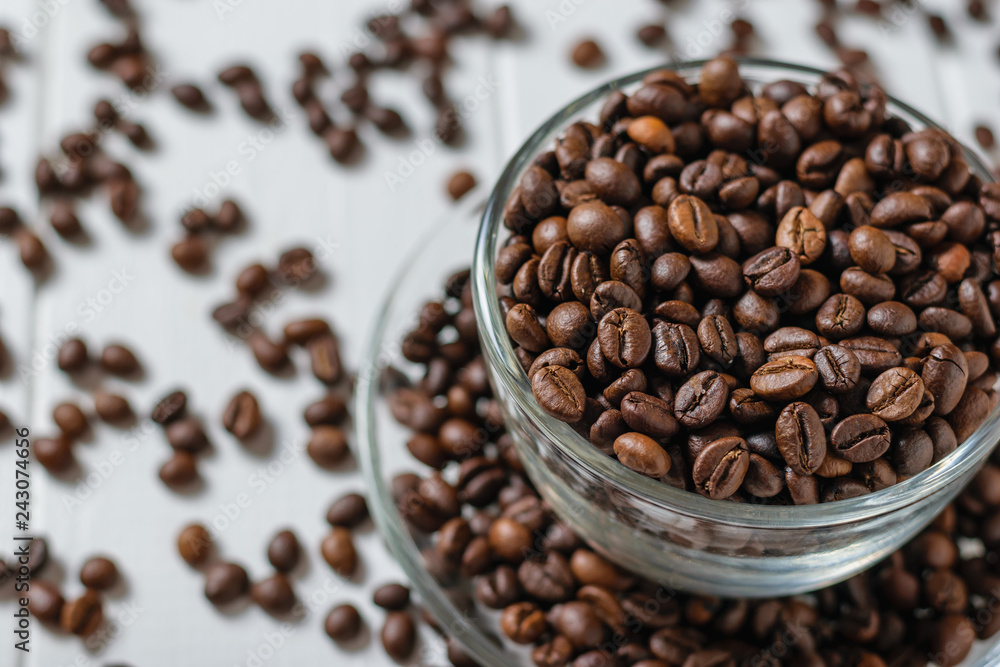 Fototapeta premium Glass bowl with saucer filled with coffee on background of scattered grains on white table.