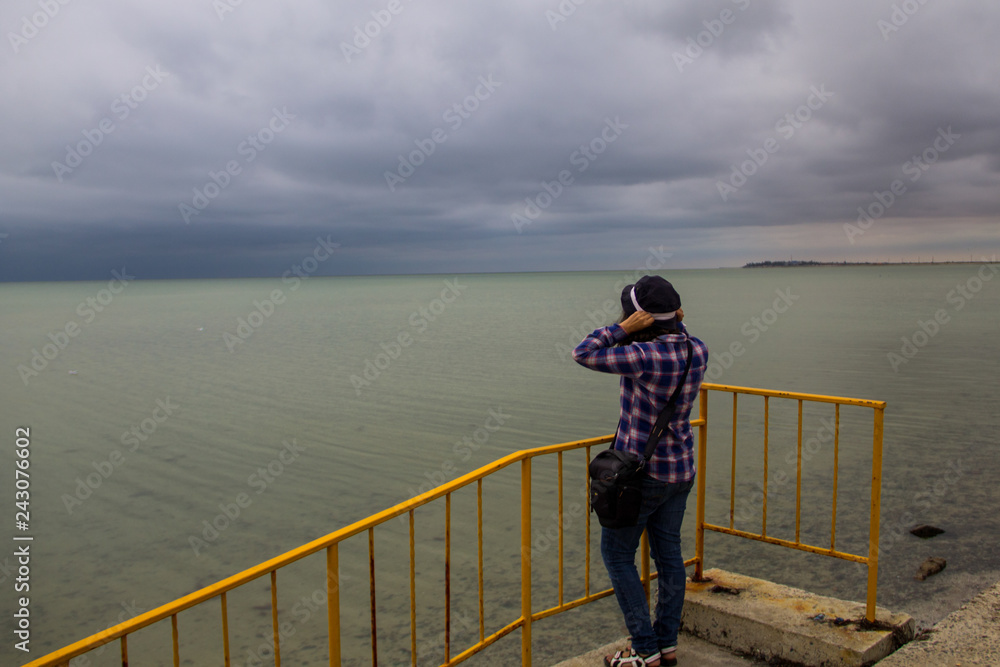 Young woman in checkered shirt standing on the beach alone and looking at sea in a gloomy gray day. Back view