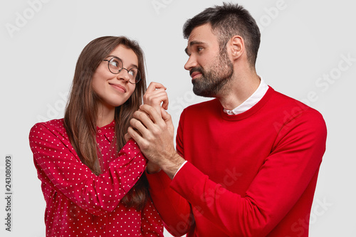 Horizontal shot of pleased woman looks at husband who has pleading expression and holds her hand, wear red clothes, have pleasant talk, isolated over white background. People, love, relations