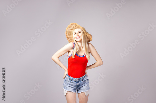 Studio shot of gorgeous young blonde woman with straight hair wearing wide  brim straw hat, tight red tank top and sexy high waisted denim shorts. Gray  isolated background, copy space, close up. Stock Photo