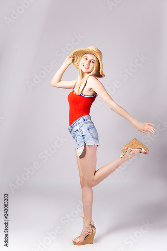 Studio shot of gorgeous young blonde woman with straight hair wearing wide brim straw hat, tight red tank top and sexy high waisted denim shorts. Gray isolated background, copy space, close up.