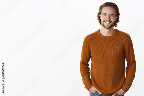 Waist-up shot of handsome stylish good-looking young boyfriend with earrings and pierced nose wearing cozy sweater holding hands in pockets and smiling happy, delighted at camera over gray wall