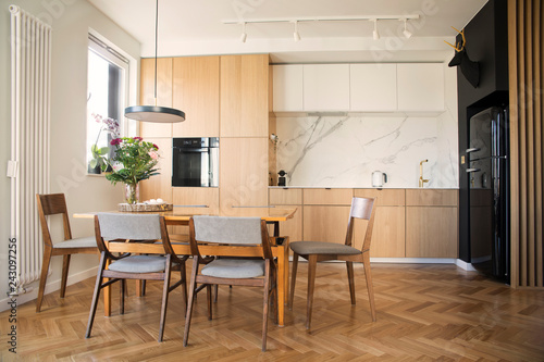 Modern scandinavian open space interior of kitchen with design furniture, black fridge, marble walls and brown wooden parquet. Elegant and floral accesories.