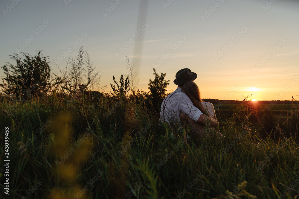 Fototapeta premium Young man and woman who are sitting together on the grass and watching sunrise photographed from behind.