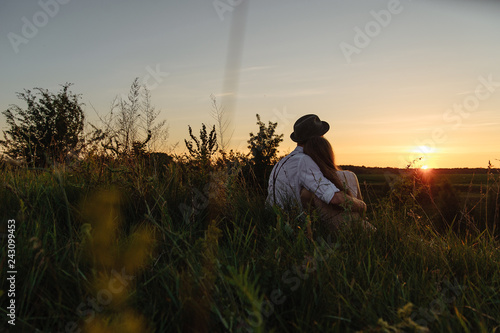 Young man and woman who are sitting together on the grass and watching sunrise photographed from behind.