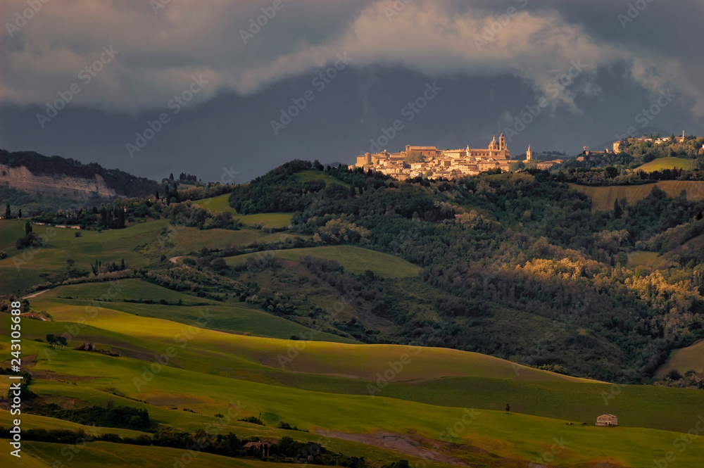 Urbino, Italy. The antique  renaissance town is illuminated by the Sun at sunrise. The meadows and the hills covered by trees stay in the shadow. The sky appears cloudy and stormy