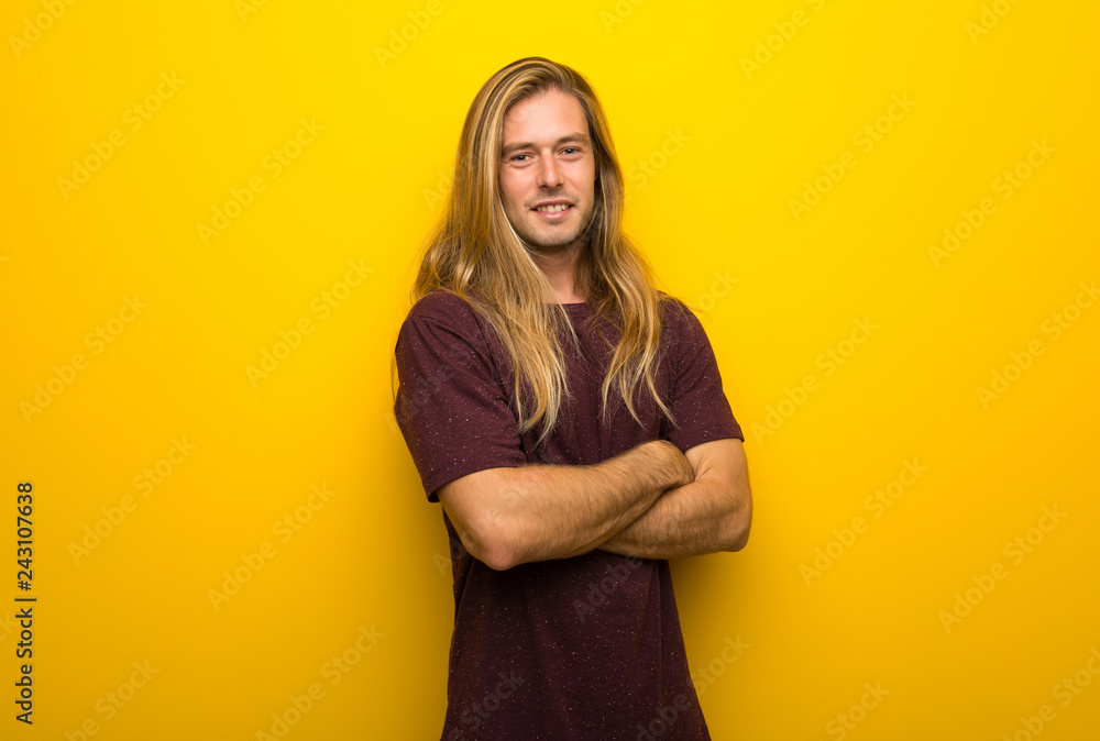 Obraz premium Blond man with long hair over yellow wall keeping the arms crossed in frontal position