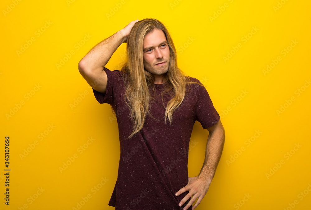 Blond man with long hair over yellow wall having doubts while scratching head Blond man with long hair over yellow wall having doubts while scratching head