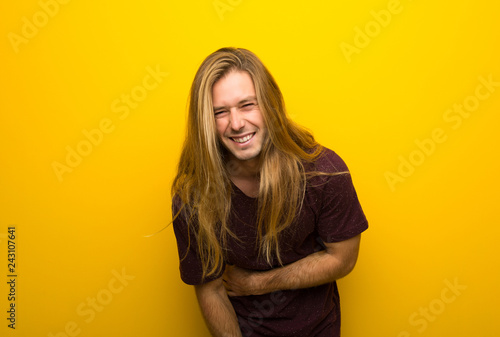 Blond man with long hair over yellow wall keeping the arms crossed while smiling
