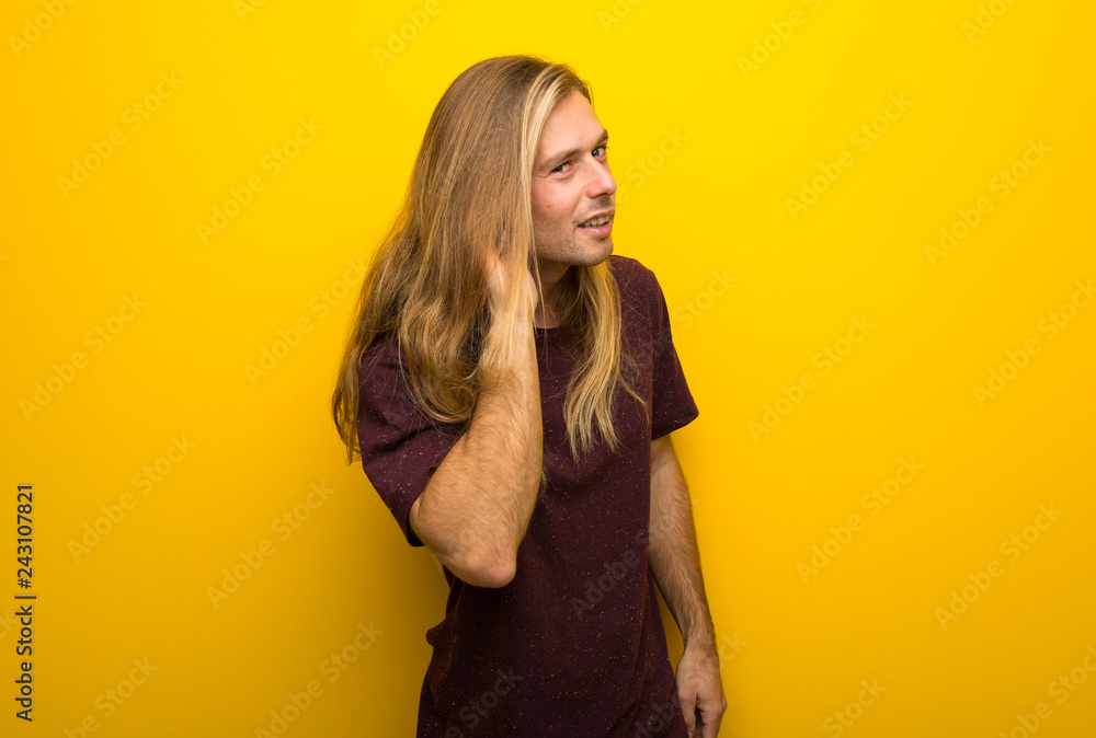 Blond man with long hair over yellow wall listening to something by putting hand on the ear Blond man with long hair over yellow wall listening to something by putting hand on the ear