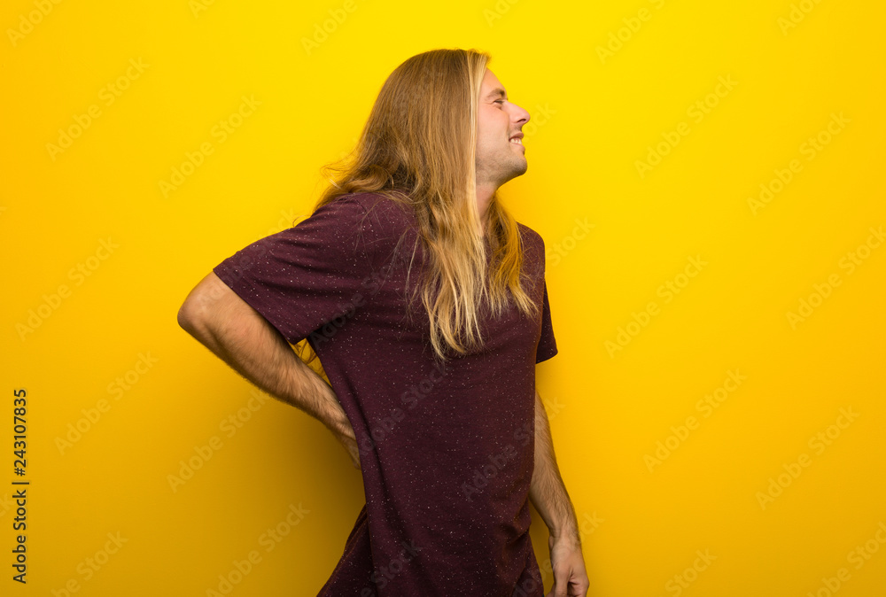 Blond man with long hair over yellow wall suffering from backache for having made an effort Blond man with long hair over yellow wall suffering from backache for having made an effort