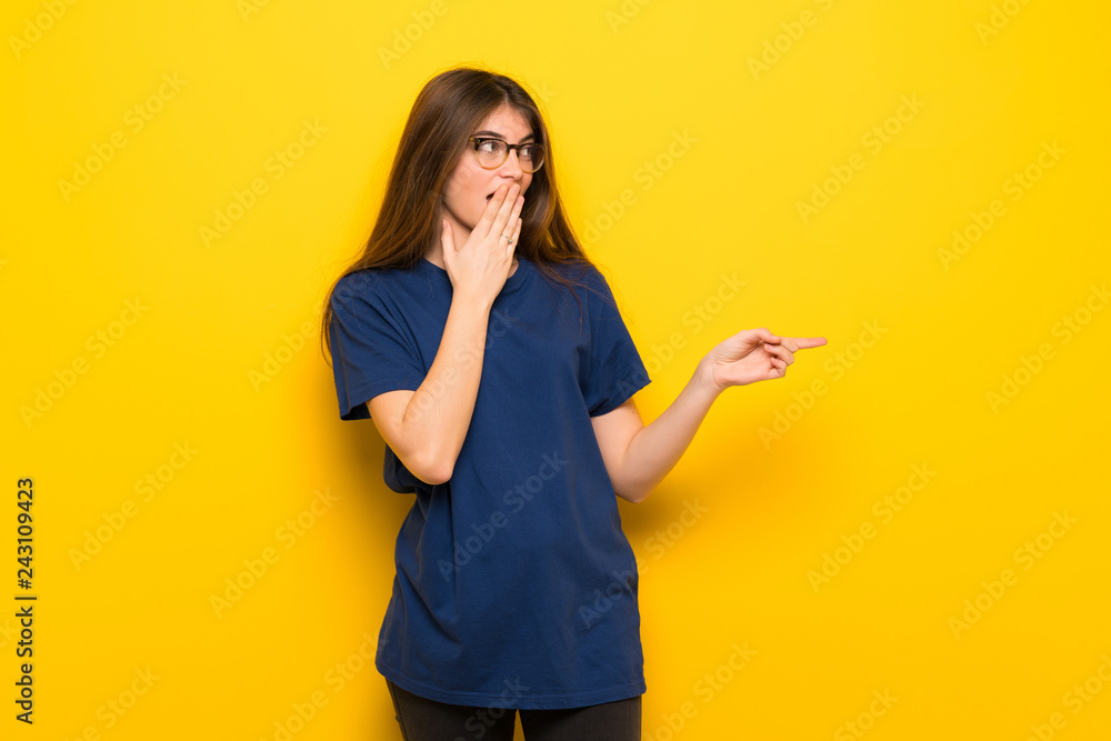 Young woman with glasses over yellow wall pointing finger to the side with a surprised face Young woman with glasses over yellow wall pointing finger to the side with a surprised face