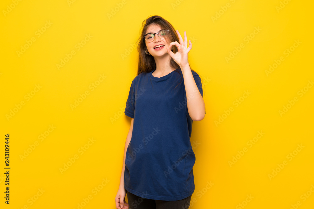 Young woman with glasses over yellow wall showing an ok sign with fingers Young woman with glasses over yellow wall showing an ok sign with fingers
