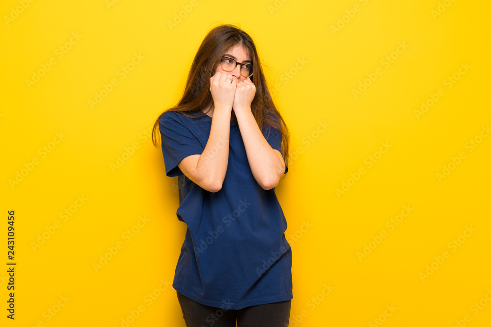 Young woman with glasses over yellow wall is a little bit nervous and scared putting hands to mouth Young woman with glasses over yellow wall is a little bit nervous and scared putting hands to mouth