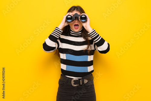 Young woman over yellow wall and looking in the distance with binoculars