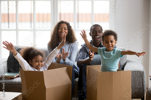 Happy african american parents and kids playing in boxes enjoy relocation into new home, excited mixed race children having fun help mom dad unpack in living room, black family on moving day portrait