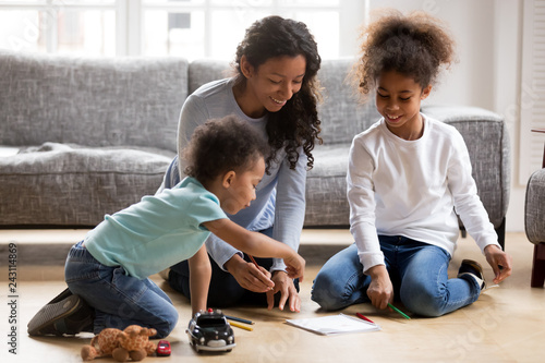 Happy black single mom and 2 mixed race kids draw with colored pencils on warm floor together, african mother baby sitter helps children son daughter playing having fun at home, creative family hobby