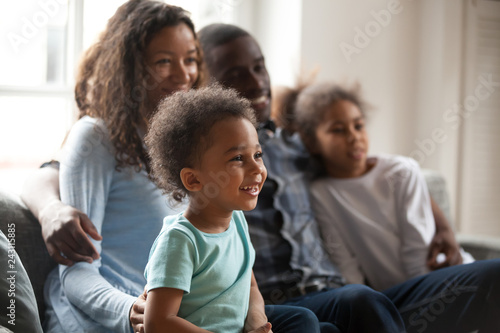 Happy black family with children sitting on couch watching tv together, african american parents embrace kids relaxing on sofa laughing enjoying funny cartoons or movie having fun on weekend at home