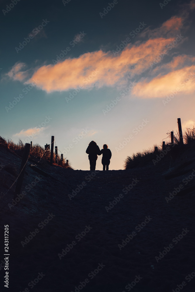 Love couple take a winter vacation walk. Silhouette of two person on beach sand dunes with sunset colorful sky tones. German Baltic Sea coastline at Fischland-Darss-Zingst in Mecklenburg