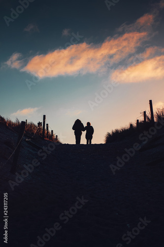 Love couple take a winter vacation walk. Silhouette of two person on beach sand dunes with sunset colorful sky tones. German Baltic Sea coastline at Fischland-Darss-Zingst in Mecklenburg