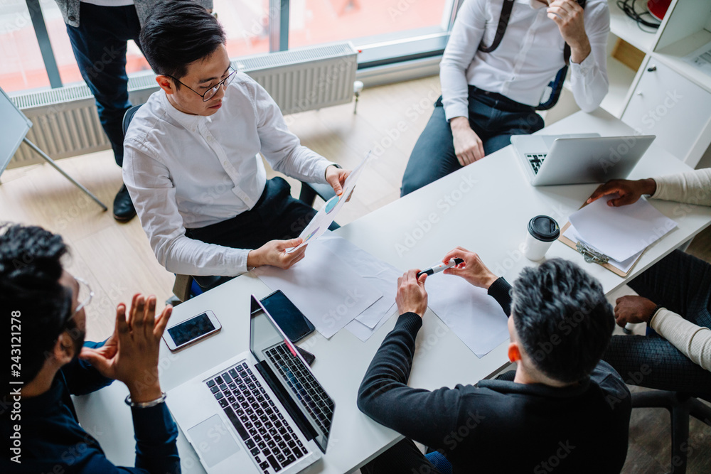 © alfa27 - Multiracial group of young people in formal wear making out business plan for future start-up project sitting at the desk. Corporate Work Concept . Top view.