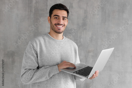 Portrait of young man in gray sweatshirt standing against textured wall, holding laptop and watching media with happy smile