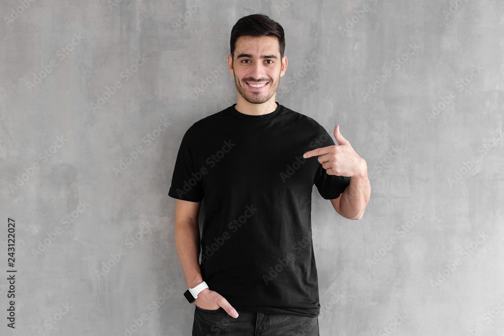 Young handsome man isolated on gray textured wall, smiling while pointing with index finger to black t-shirt, copyspace for advertising - obrazy, fototapety, plakaty 