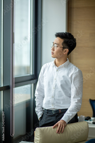Asian office employee in white shirt standing near the window and looking at the cityscape