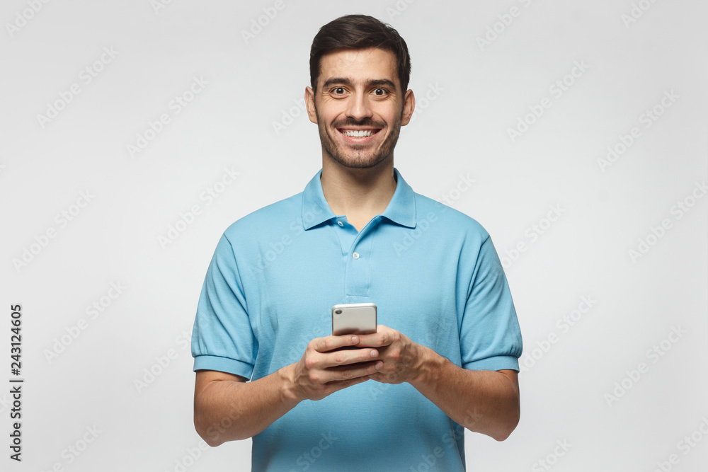 Young man standing isolated on gray background, holding smartphone, looking at camera and smiling nicely Young man standing isolated on gray background, holding smartphone, looking at camera and smiling nicely