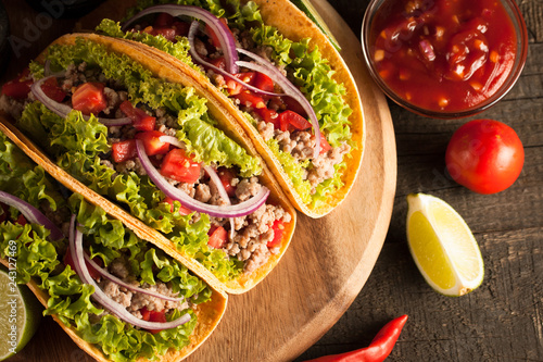 Photo of Mexican tacos with ground beef, onion, tomatoes, chili, red sauce, lettuce and lime on wooden background. Spicy and fast food concept.