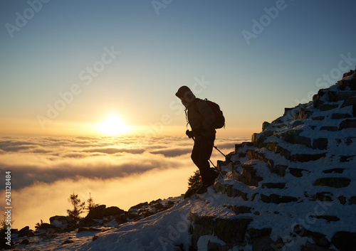 Tourist climber with backpack hiking on rocky mountain steep slope covered with first snow on copy space background of foggy valley filled with white puffy clouds, raising sun and blue sky at dawn.