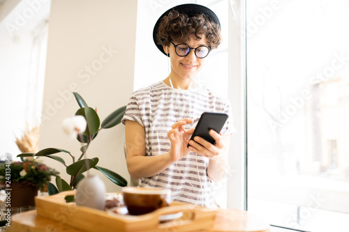 Smiling attractive modern young woman in hat and stripped tshirt sitting on window sill and listening to music in earphones while using smartphone in cafe