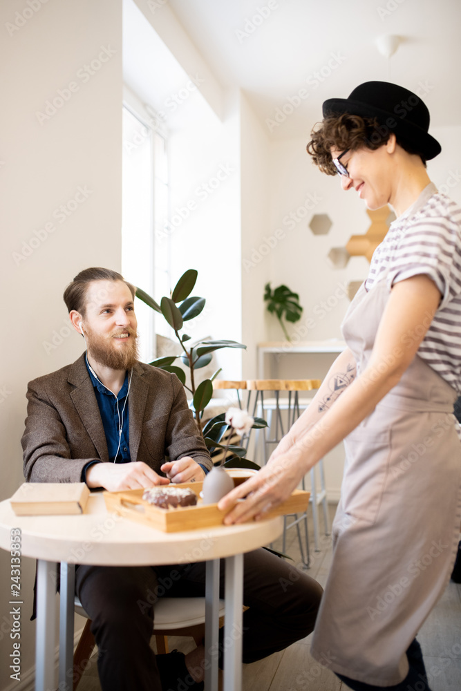 Smiling friendly waitress in hat bringing order to customer in modern coffee shop and putting tray on table, cheerful bearded man in earphones smiling at waitress Smiling friendly waitress in hat bringing order to customer in modern coffee shop and putting tray on table, cheerful bearded man in earphones smiling at waitress