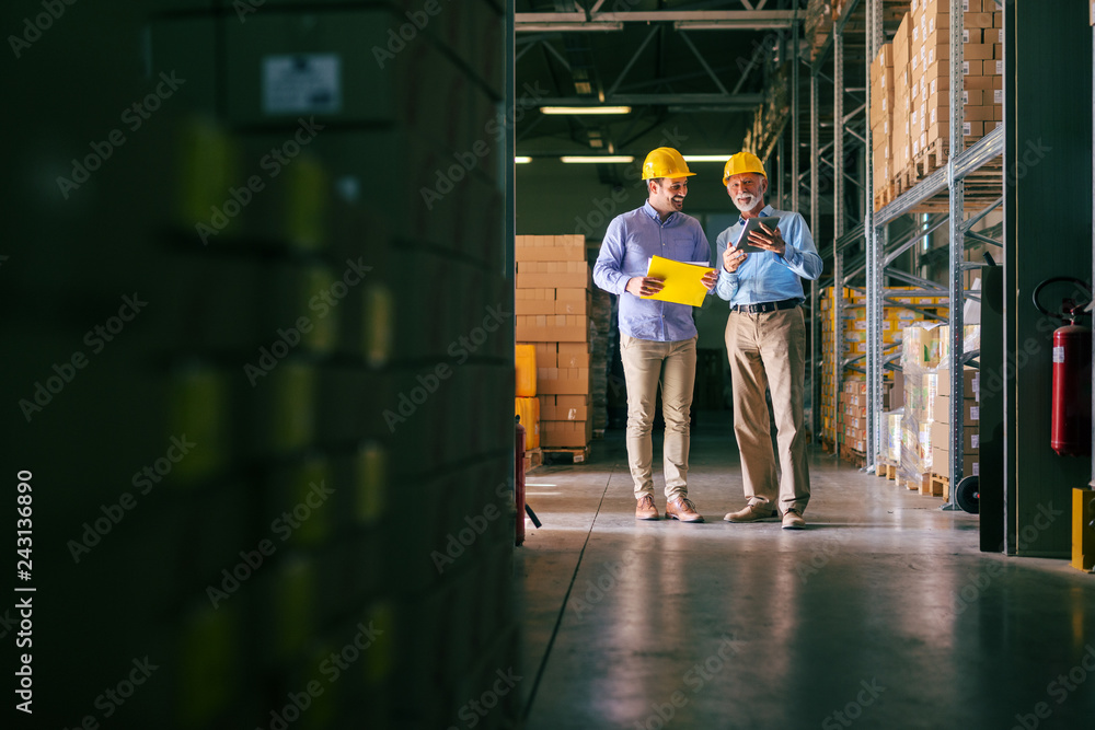 Two bearded businessmen with helmets on head comparing documents while standing in warehouse.