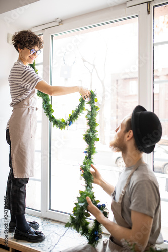 Positive young colleagues in aprons hanging garland on window together in cafe: hipster man giving Christmas tinsel to lady on window-sill
