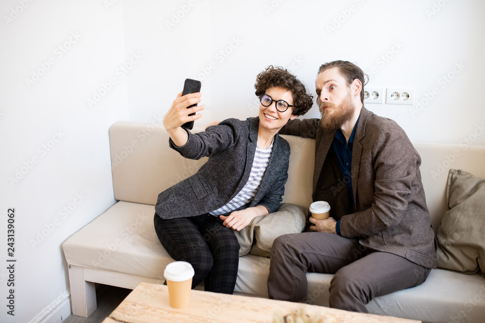 Smiling attractive curly-haired lady in glasses sitting on sofa and photographing with funny friend in modern coffee shop Smiling attractive curly-haired lady in glasses sitting on sofa and photographing with funny friend in modern coffee shop