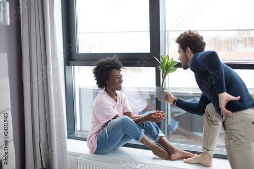 Loving happy interracial couple sitting on the sill and looking at the large window in their hired flat. Caucasian guy wears a dark poolover, african girl wears pink shirt.