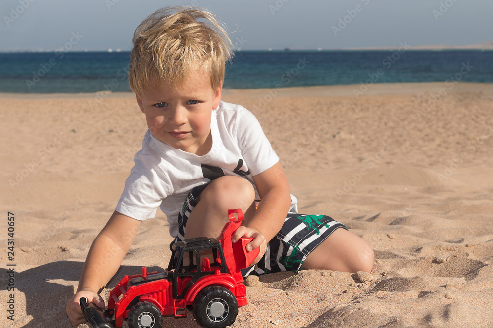 Caucasian child boy playing toy red tractor, excavator on a sandy beach at sunny day. Caucasian child boy playing toy red tractor, excavator on a sandy beach at sunny day.