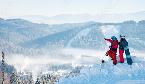 Two snowboarders - man and woman enjoying beautiful natural landscape, resting on top of the mountain on a sunny winter day at ski resort. Man pointing to the background copyspace lifestyle recreation