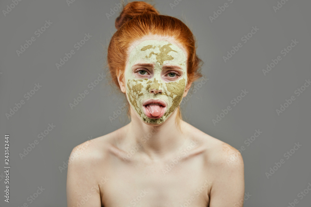 young woman with green mask feels bored. close up photo. nervous girl with mud mask showing tongue