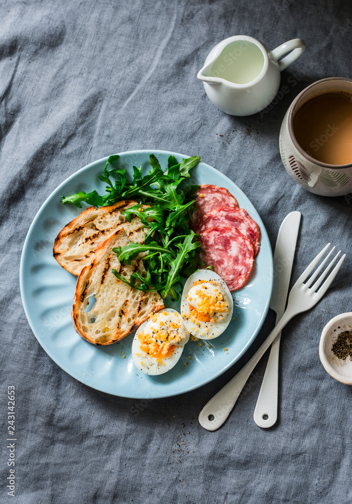 Delicious breakfast or snack - salami sausage, boiled egg, arugula, grilled bread and coffee on a gray background, top view