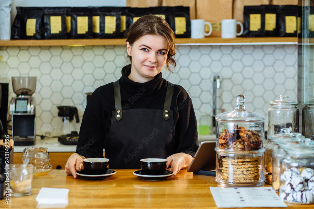 Female caucasian barista with two cups of cappuccino behind counter in modern coffee shop. Female caucasian barista with two cups of cappuccino behind counter in modern coffee shop.