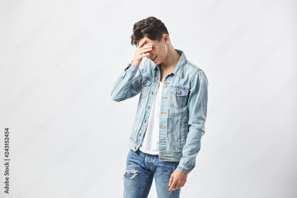 Sad dark-haired guy dressed in a white t-shirt, jeans and a denim jacket holds his hand on the face on the white background in the studio Sad dark-haired guy dressed in a white t-shirt, jeans and a denim jacket holds his hand on the face on the white background in the studio