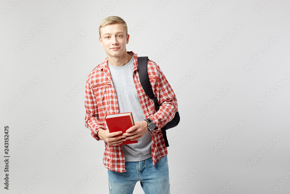 Young blond guy with black backpack on his shoulder dressed in a white t-shirt, red checkered shirt and jeans holds books in his hands on the white background in the studio Young blond guy with black backpack on his shoulder dressed in a white t-shirt, red checkered shirt and jeans holds books in his hands on the white background in the studio