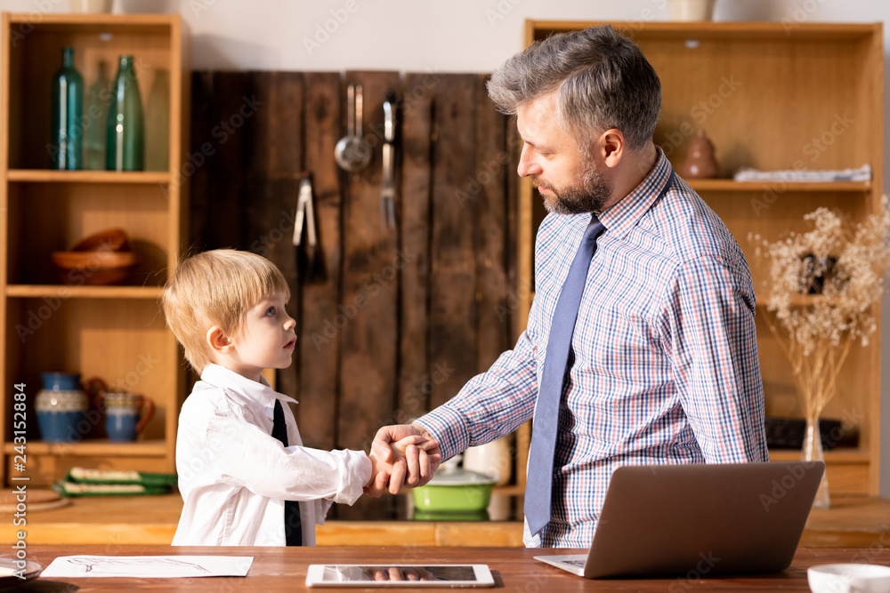 Serious confident handsome bearded father in shirt and tie standing in kitchen and making handshake with little son in formal shirt Serious confident handsome bearded father in shirt and tie standing in kitchen and making handshake with little son in formal shirt