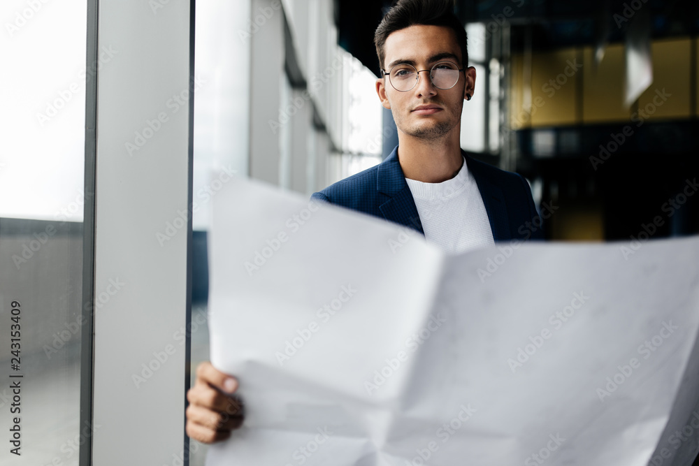 Fototapeta premium Architect in stylish clothes holds sheet with drawing in his hand and talks by phone on the background of a modern glass multistory building